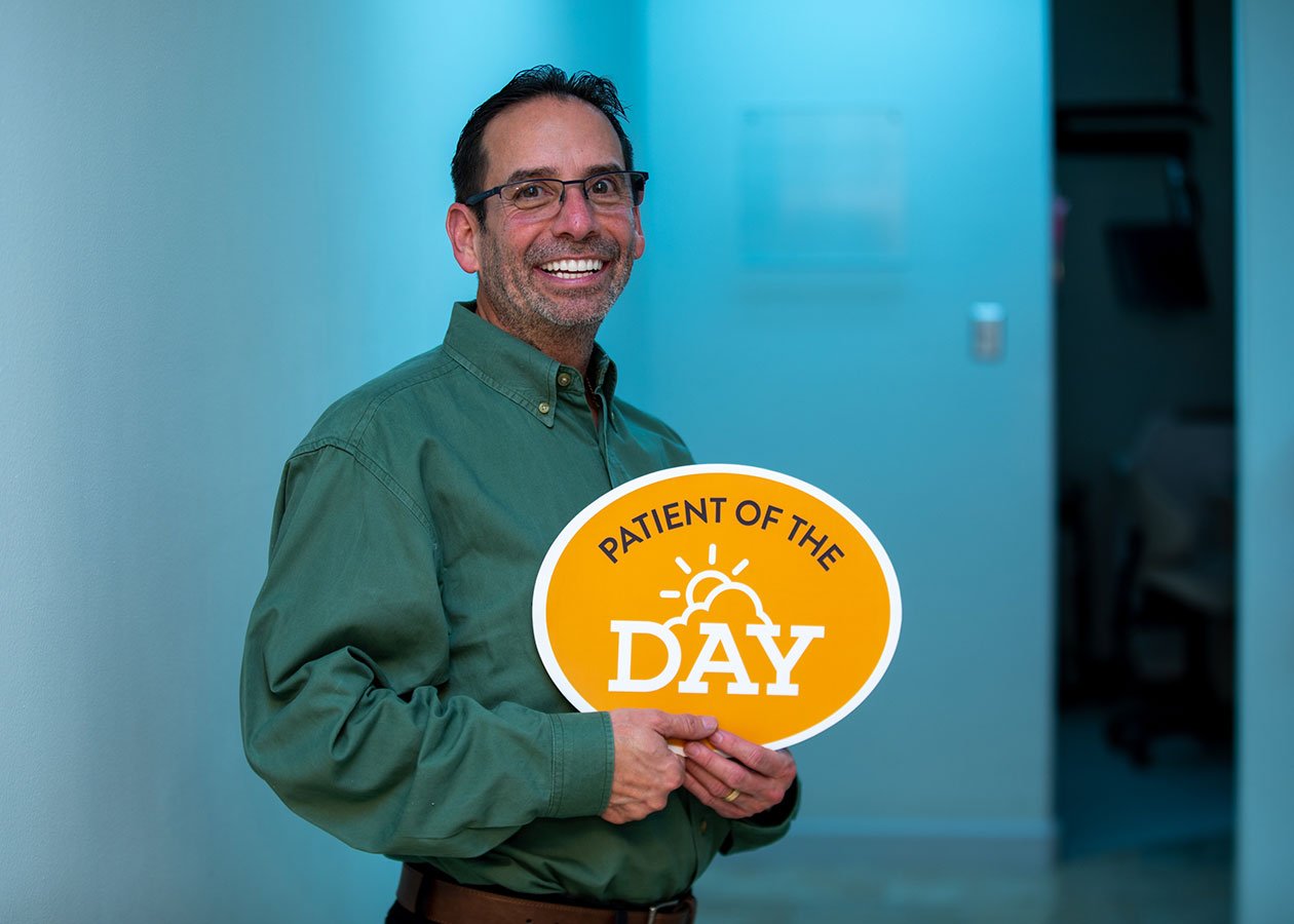A man with glasses and a green shirt smiling while holding a sign that says 'Patient of the Day' with a sun and sunrise graphic.