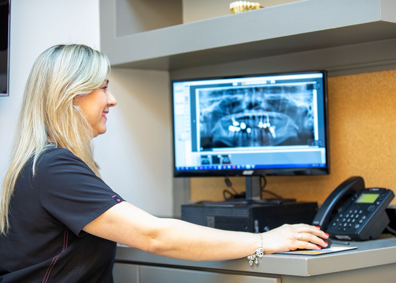 A woman is sitting at a desk, looking at a computer screen displaying a dental X-ray. She is wearing a black uniform and has blonde hair, with her right arm extended toward a telephone.