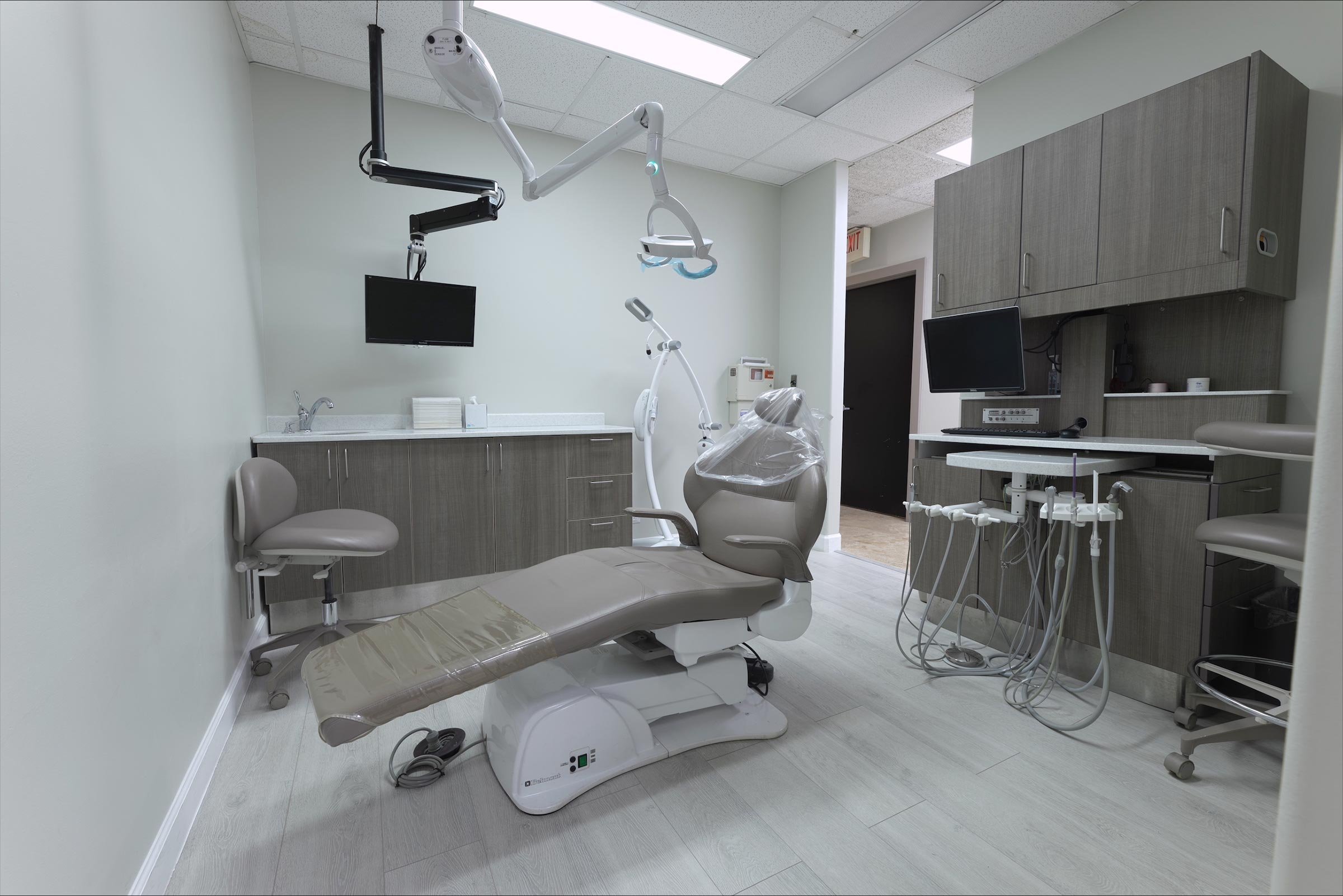 Empty dental examination room with dental chair covered in plastic, overhead lights, monitor, and various dental equipment and cabinets.