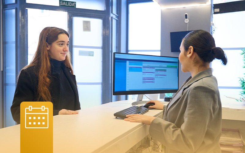 A woman at the reception desk at the dentist. The attendant has a calendar pulled up to schedule the woman for an appointment. There is an icon for a calendar in the bottom left corner of the image.