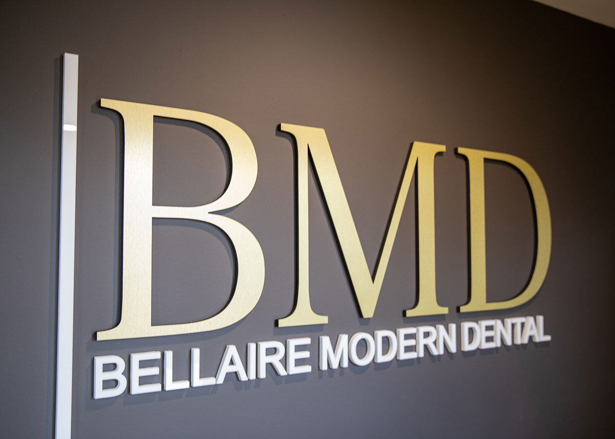 Sign for Bellaire Modern Dental with large gold letters BMD and smaller white letters below reading Bellaire Modern Dental, mounted on a gray wall.