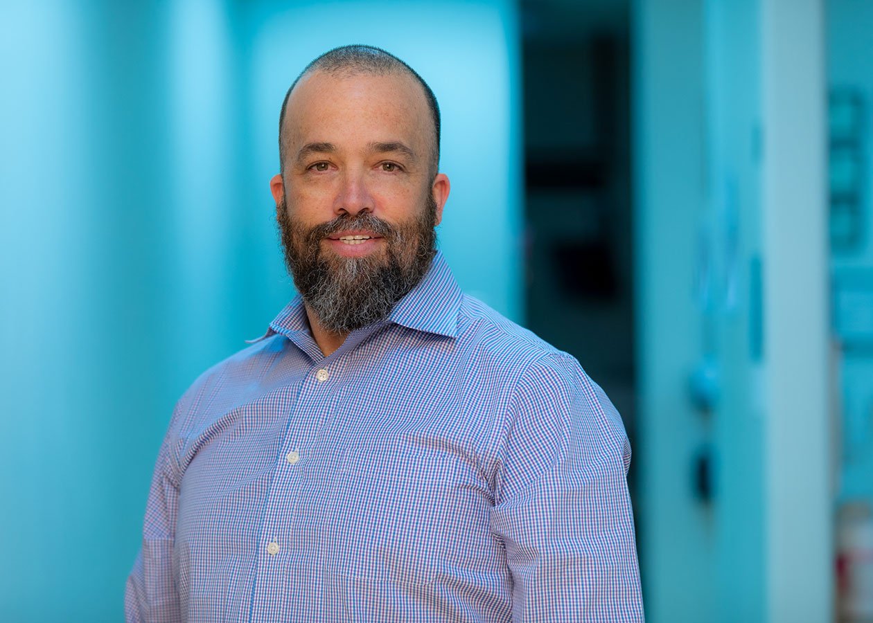 A man with a beard and short hair, wearing a checkered dress shirt, standing outdoors near a light-colored building.