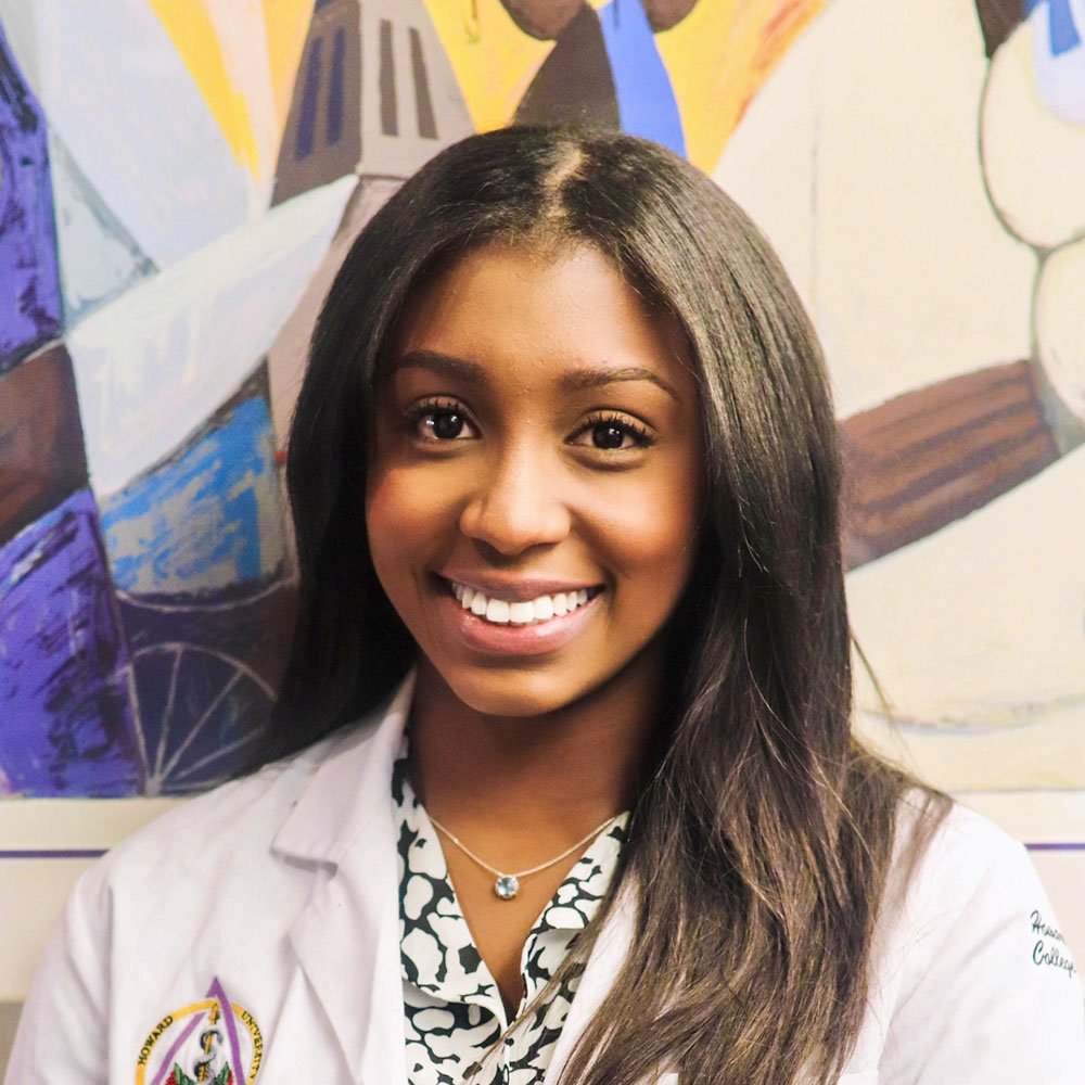 Young woman smiling in front of a colorful artwork, wearing a white coat and a black and white patterned shirt, with a necklace.